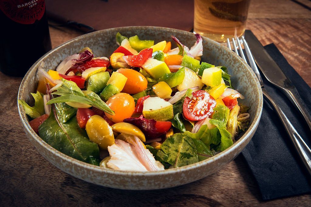 Colorful salad in a gray bowl with tomatoes, avocado, and peppers; cutlery and drink in background.
