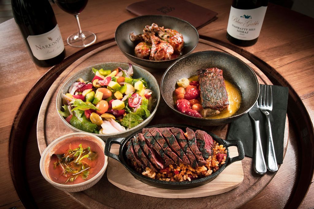 A well-lit table with a gourmet meal and wine, including steak, salad, and roasted vegetables.