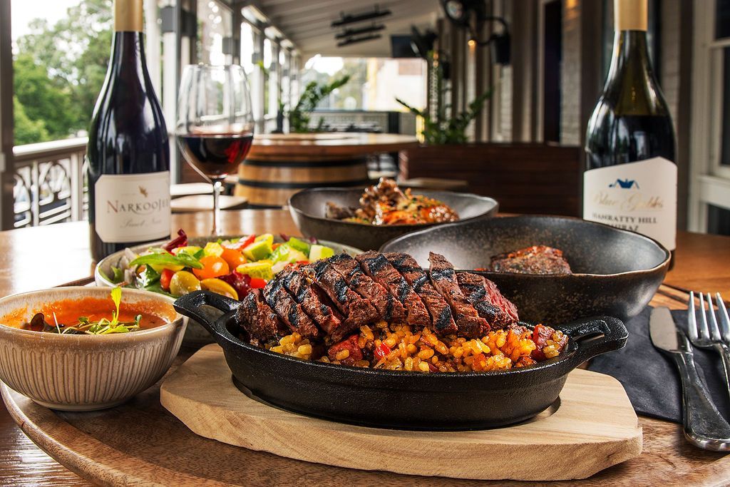 Meal set on a wooden table with steak, rice, salad, and wine bottles. Outdoor setting.