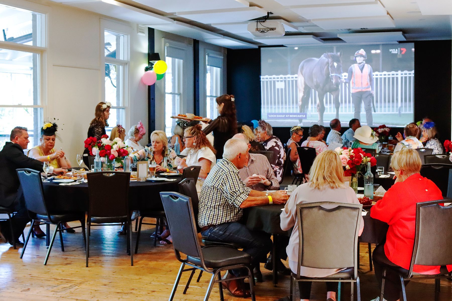 People watch a horse race on a screen in a room with tables, hats, and flowers.