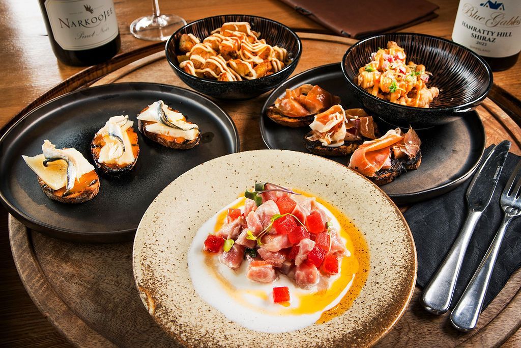 Seafood dishes on plates, arranged on a wooden tray. Two bottles of wine and silverware are visible.