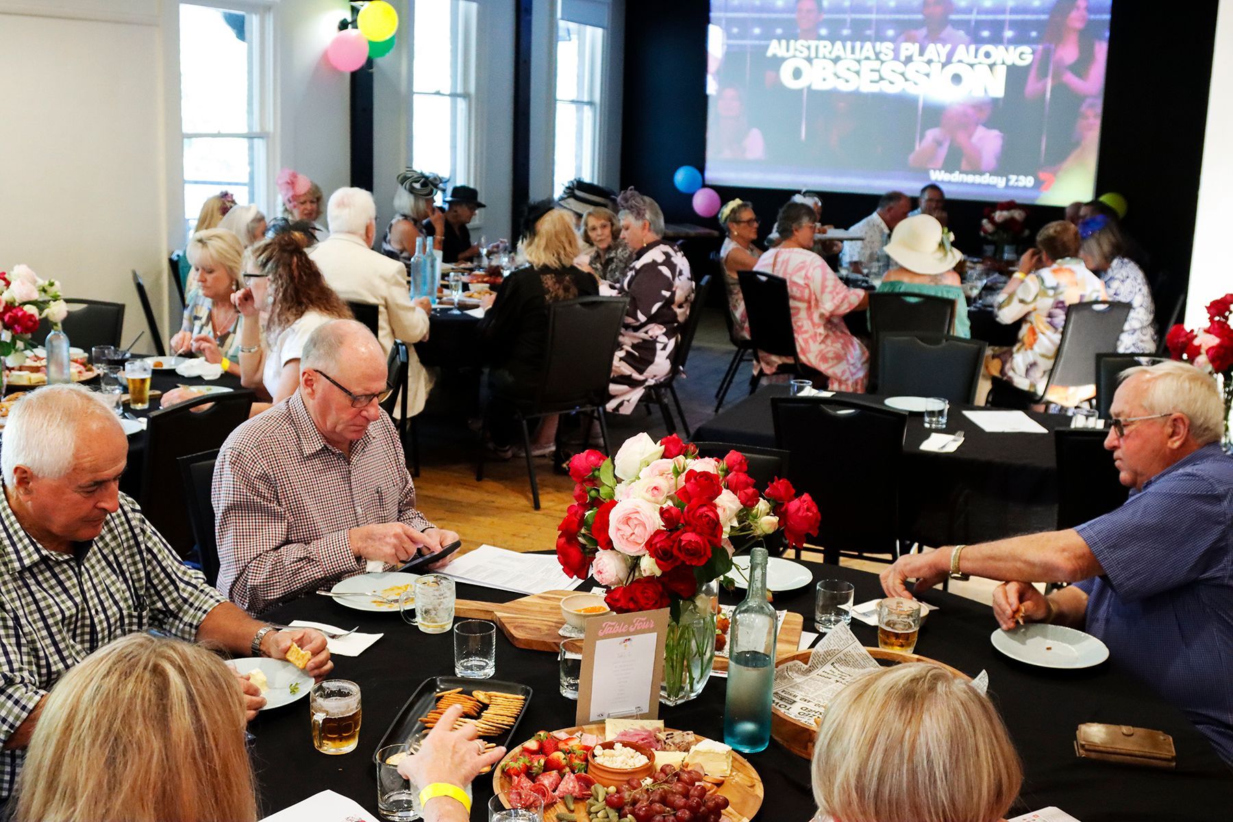 People at tables, eating at an event with a screen displaying a title. Many wear hats, flowers on tables.