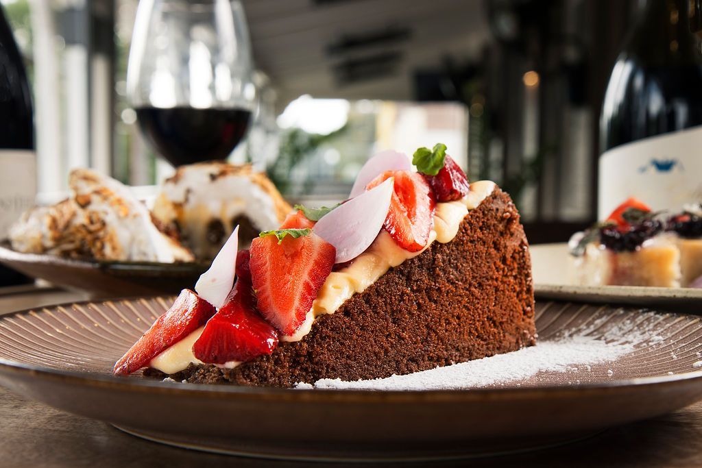 Slice of chocolate cake topped with strawberries on a brown plate with other food items and wine bottles in the background.