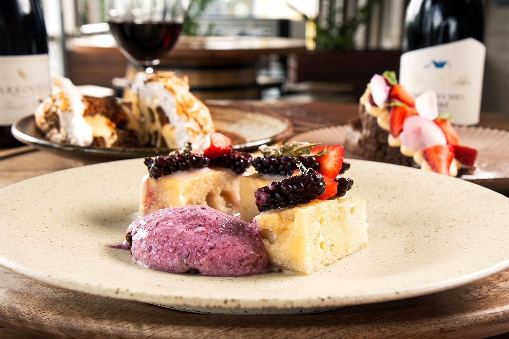 Dessert plate with cake, purple mousse, blackberries, and strawberries; wine bottles and glass in background.