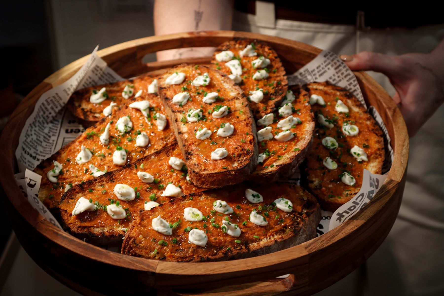 Wooden tray holding toasted bread slices topped with red sauce, white cheese, and chives.