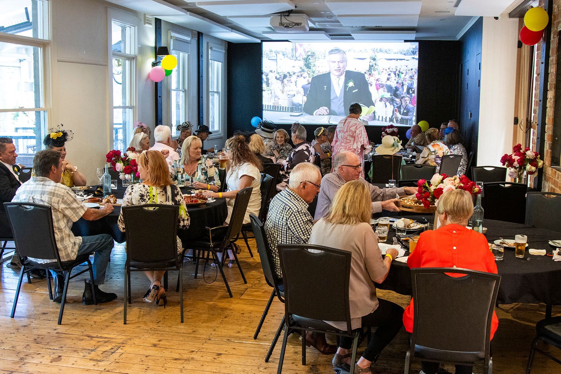 People seated at tables in a room, watching a presentation on a screen. Black table cloths, balloons, and flowers.