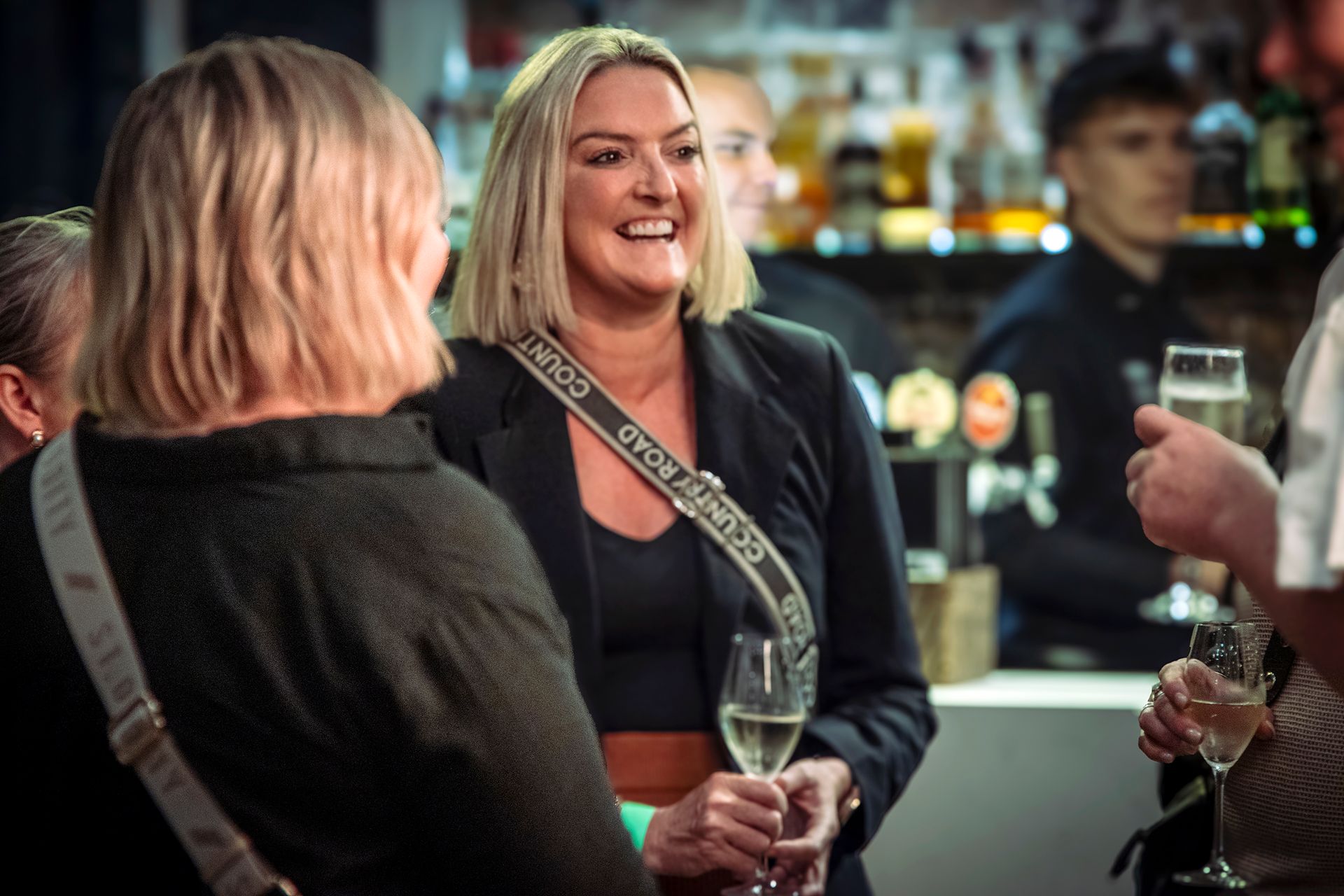 Woman smiling, holding a glass of champagne, talking to others in a bar setting.