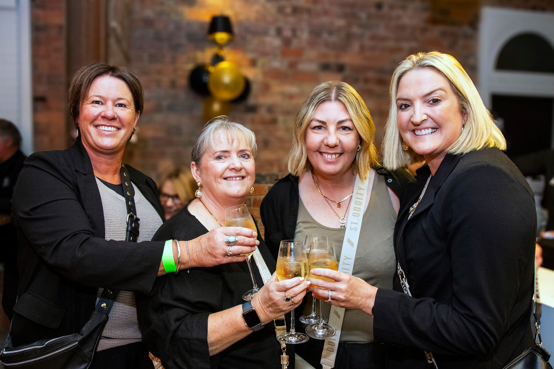 Four women toasting champagne glasses at an event, smiling, brick wall backdrop.
