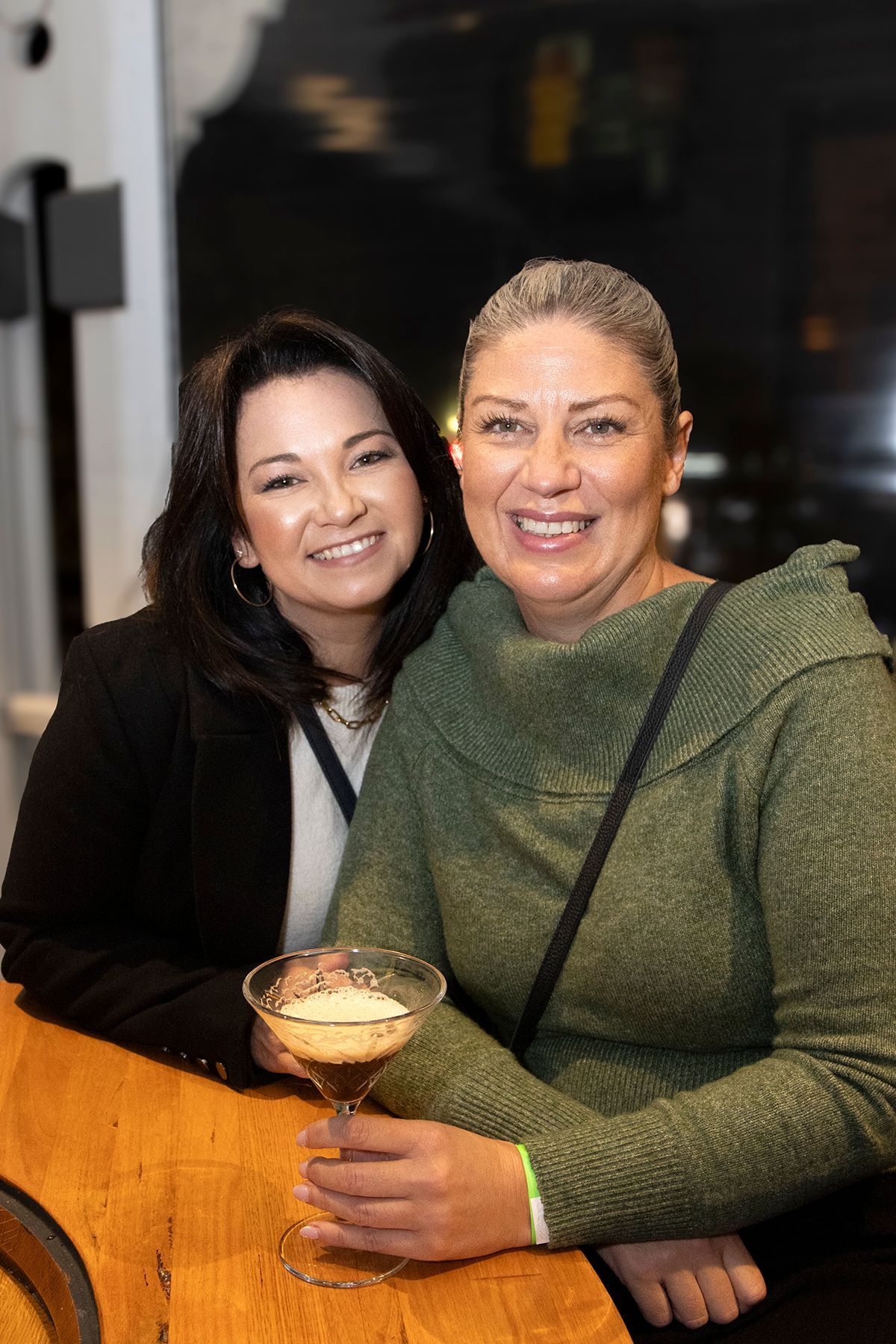 Two women smile, one holding a cocktail with foam, seated at a wooden table in a dimly lit setting.