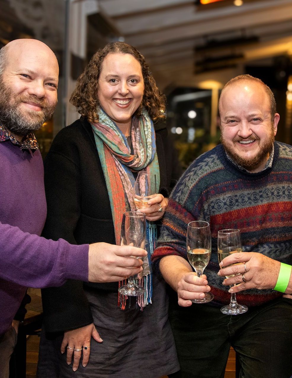 Three people toasting with champagne flutes indoors, smiling. One in purple, one in black, and one in a patterned sweater.