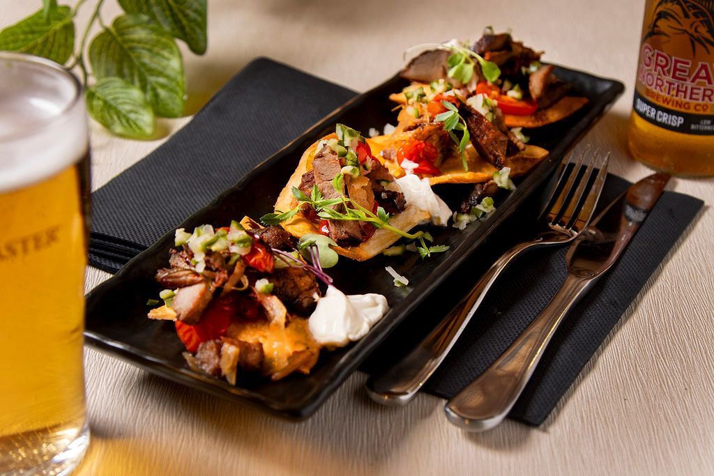 Tostadas topped with meat, vegetables, and sauce on a black plate, with beer, cutlery, and napkin.