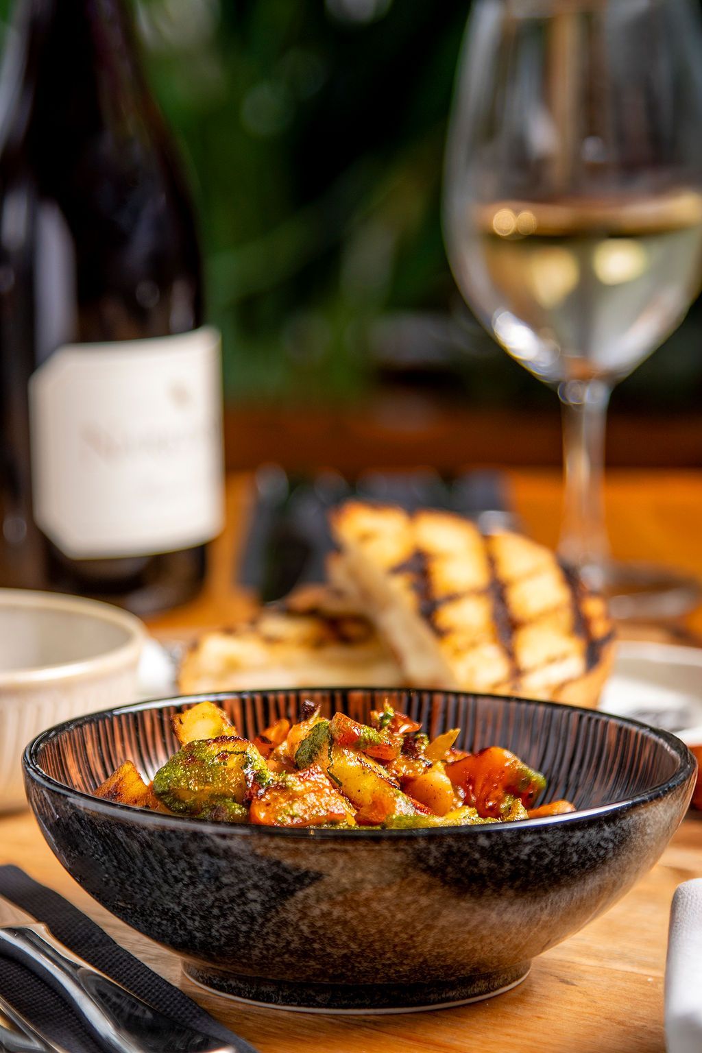 Bowl of colorful vegetables with grilled bread, wine, and a bottle on a wooden table.