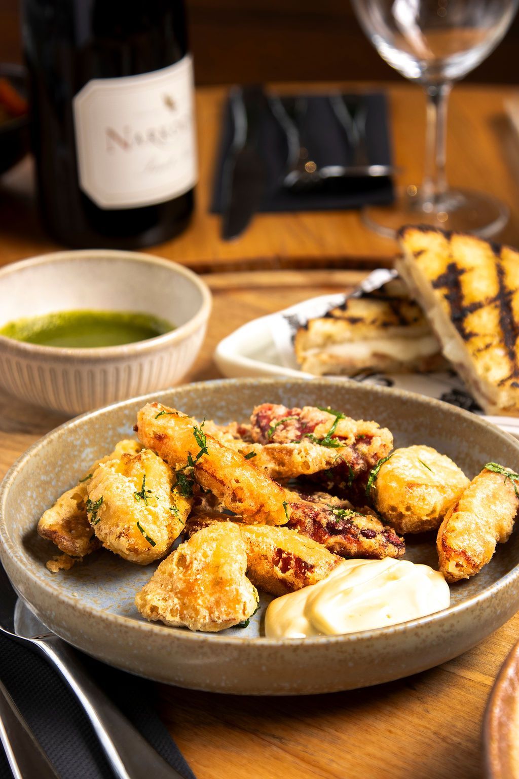 Fried seafood dish with dipping sauces, grilled bread, wine bottle on a wooden table.