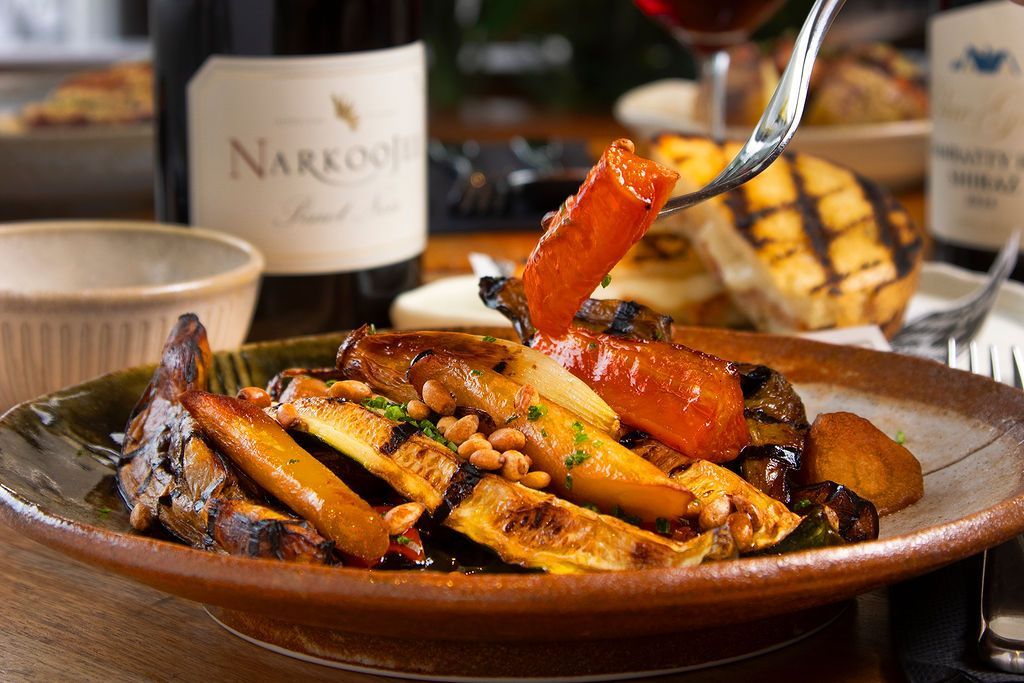 Roasted vegetables with pine nuts, carrots, and eggplant on a plate, near wine bottles.