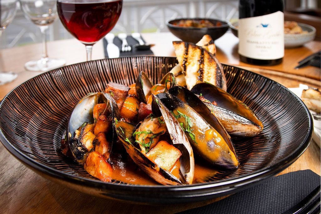 Mussels in a dark bowl, served with bread and red wine, on a wooden table.