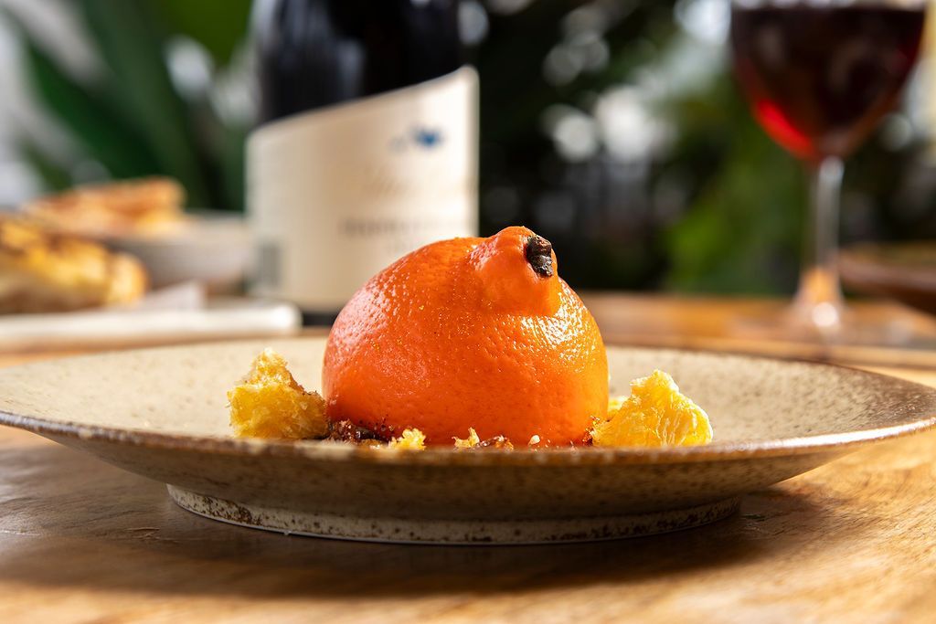Dessert on plate: orange sphere, crumbs, and chunks with wine in glass and bottle in the background.