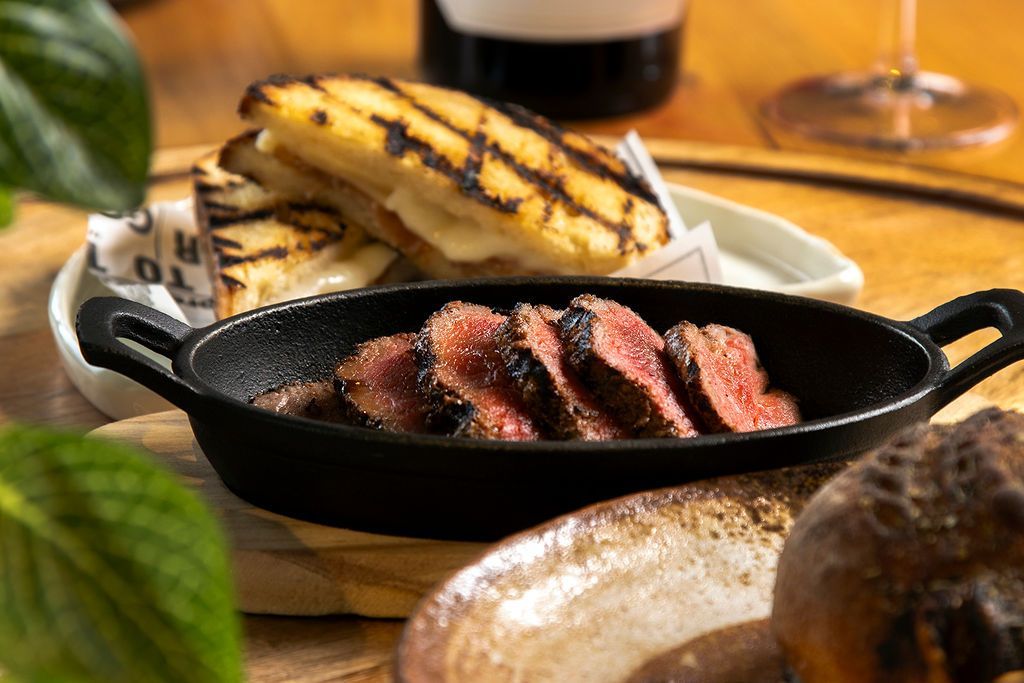 Sliced steak in a cast iron dish, grilled sandwich, wine glass, and wooden table.