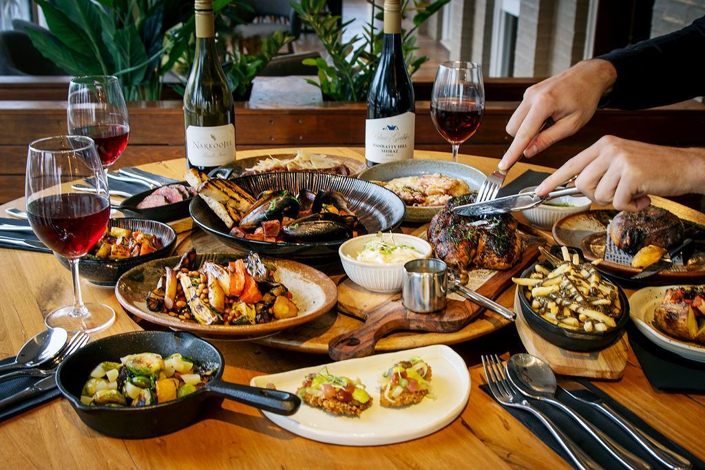 A table laden with various dishes and wine, with hands reaching for food; restaurant setting.