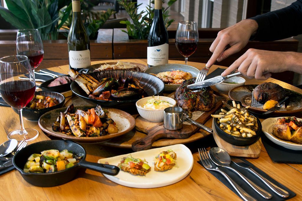 A table laden with food, including steaks, vegetables, and wine. People eating with silverware at a restaurant.