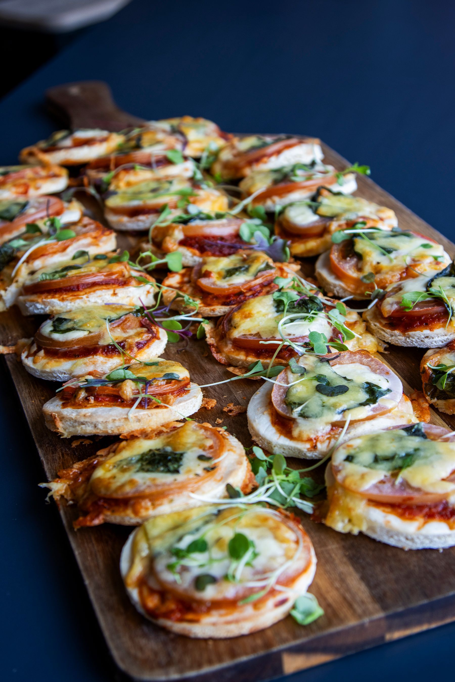 Mini pizzas on a wooden board, topped with cheese, tomato, and greens, against a blue background.