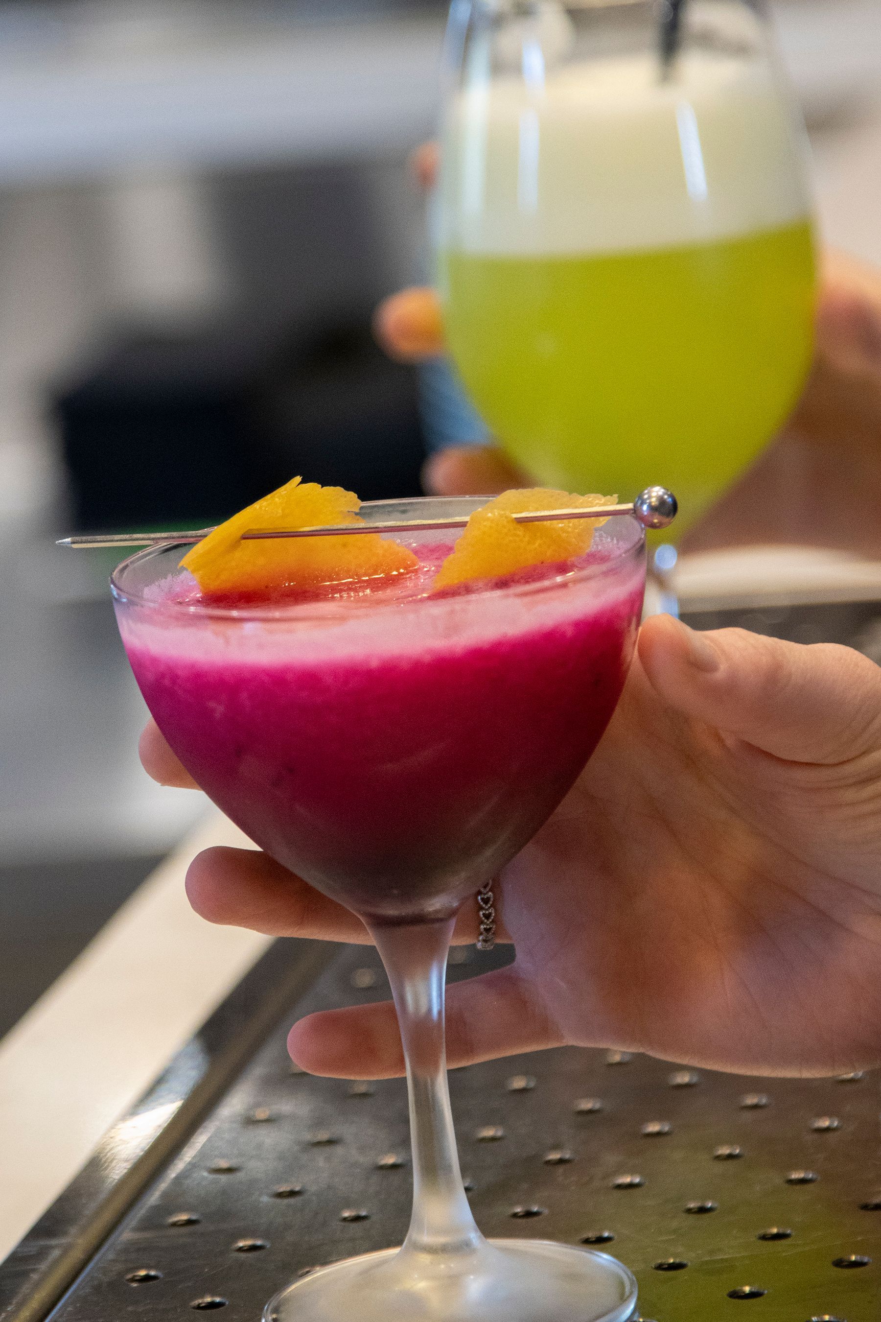 Cocktails: a magenta one with orange garnish, and a lime green one, held by hands at a bar.
