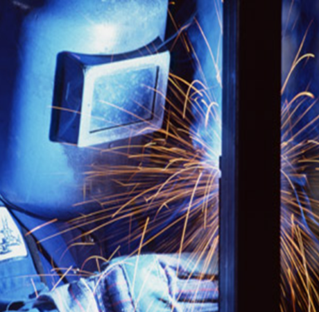 A welder wearing a protective mask works on metal, surrounded by bright orange sparks in a dark workspace.