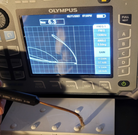 An Olympus eddy current flaw detector displays a test signal while a probe scans a white surface with raised dots.
