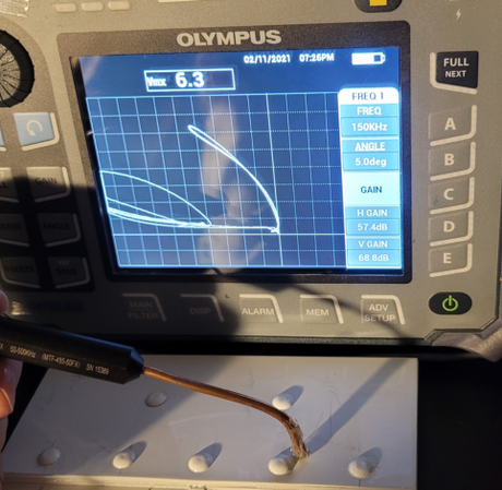 An Olympus eddy current flaw detector displays a test signal while a probe scans a white surface with raised dots.