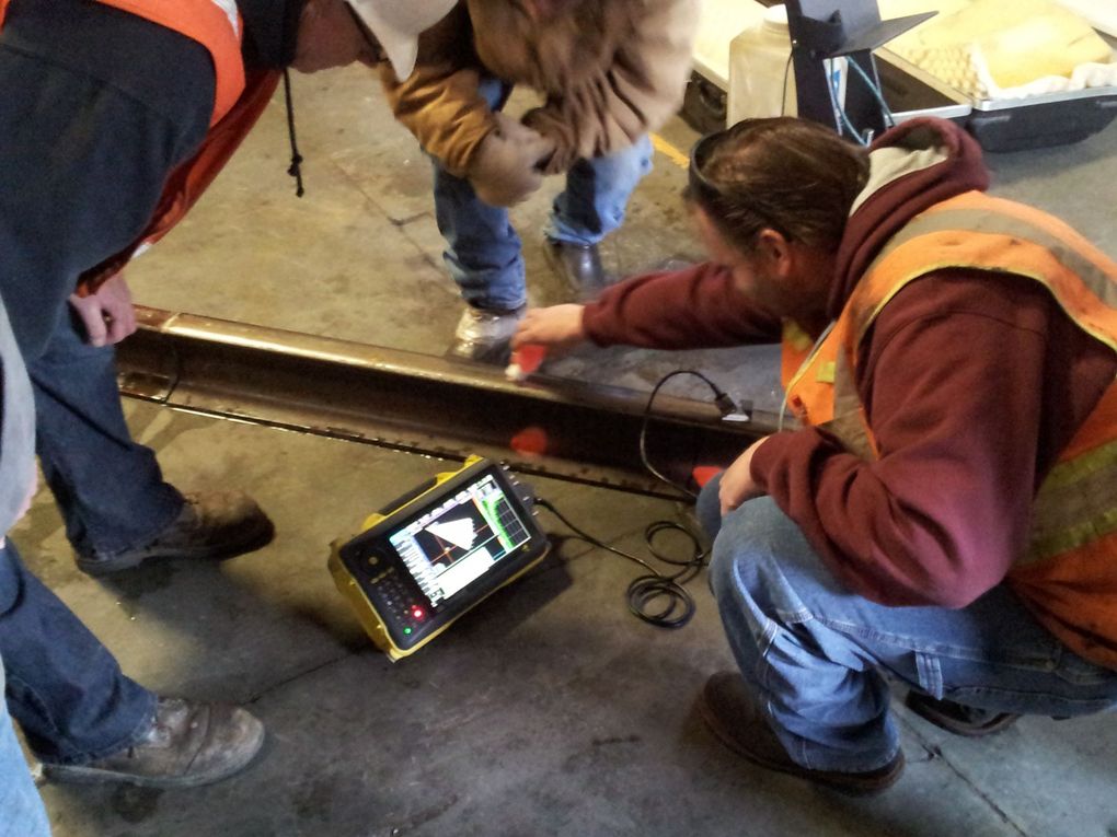 Technicians use an ultrasonic testing device to inspect a steel rail on a concrete floor in an industrial setting.