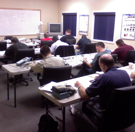 A group of people sit at tables in a classroom, working on papers and electronic devices in front of a whiteboard.