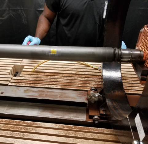 A person wearing blue gloves works on a metal rod mounted on a machine above a surface made of wooden slats.
