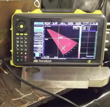 A Sonatest ultrasonic testing device displays a red sector scan on its screen next to a metal calibration block.