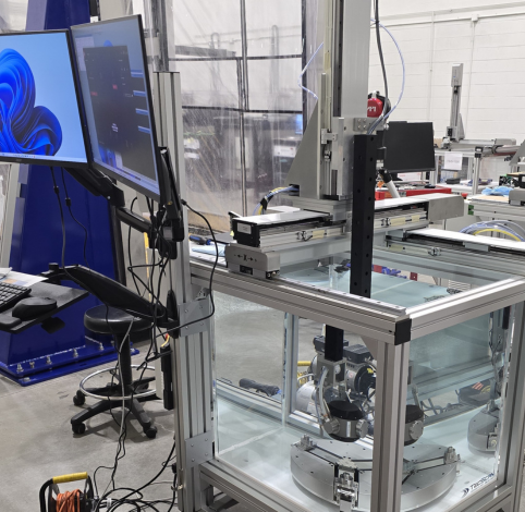 A robotic workstation in a lab features a multi-axis arm over a clear enclosure, with computer monitors on a side stand.