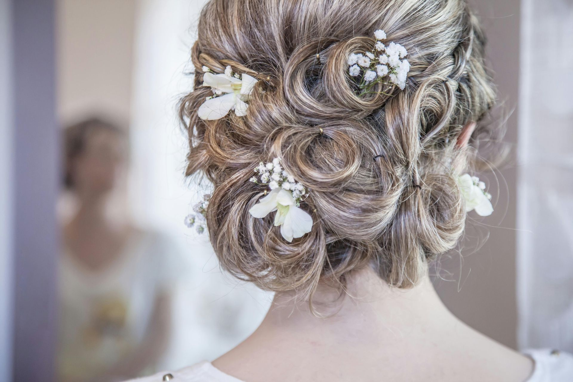 Updo hairstyle with intricate curls, light highlights, and small white floral accents, viewed from behind.