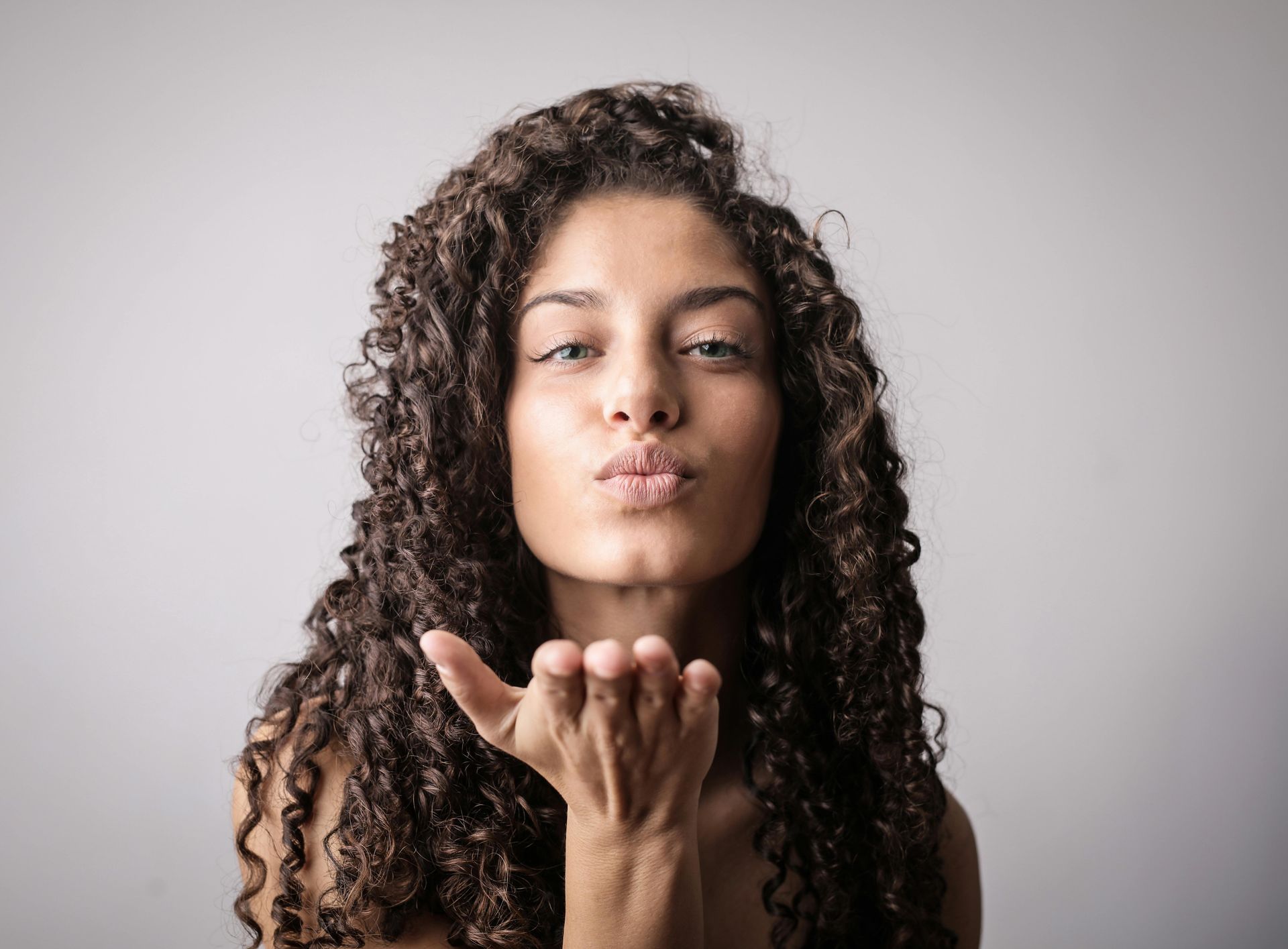 Woman blowing a kiss; curly hair, light skin, hand up, against a gray background.
