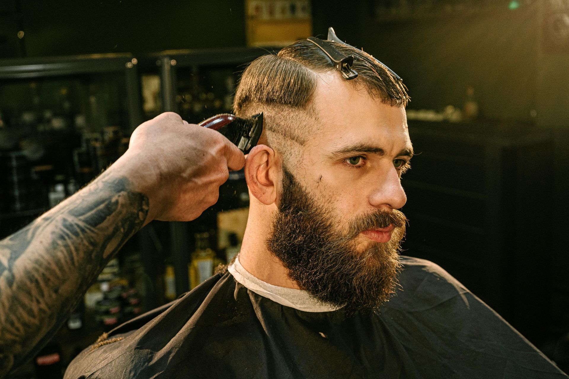 Barber cutting a man's hair with clippers. Man has a full beard and is wearing a barber's cape.