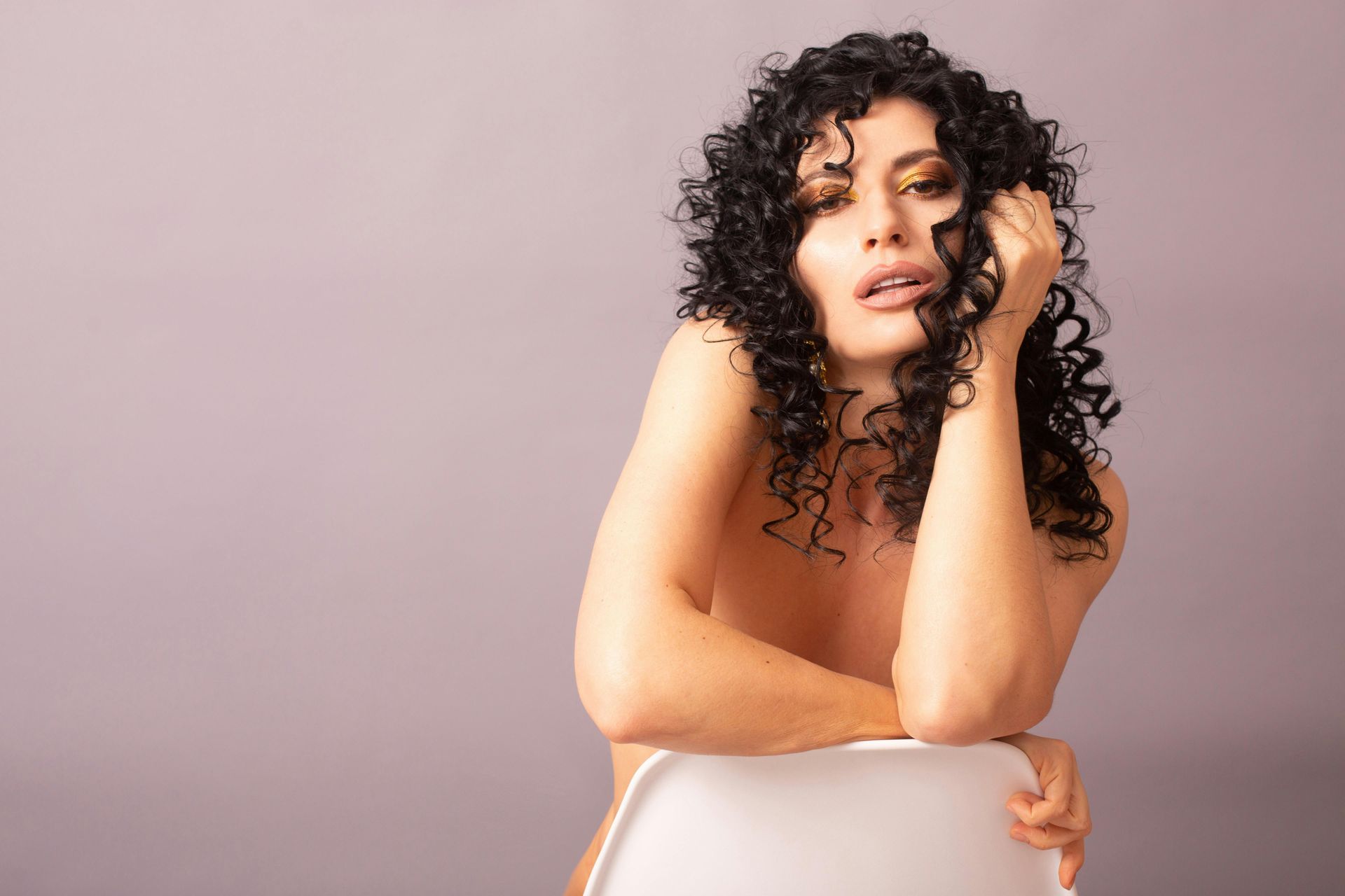 Woman with curly dark hair leans on a white chair, looking at the viewer. Neutral background.