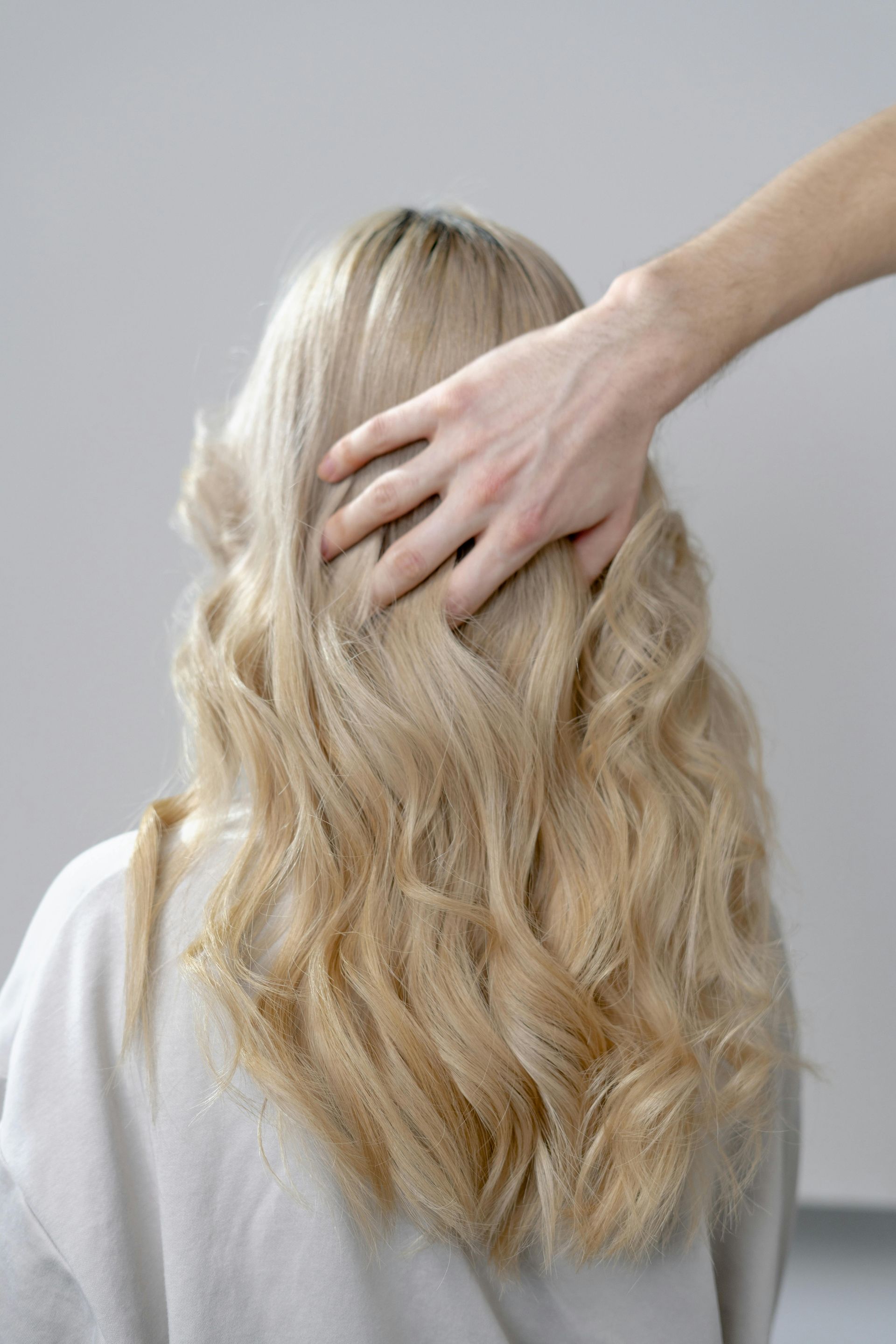 Person's back with long wavy blonde hair being touched by a hand; light background.