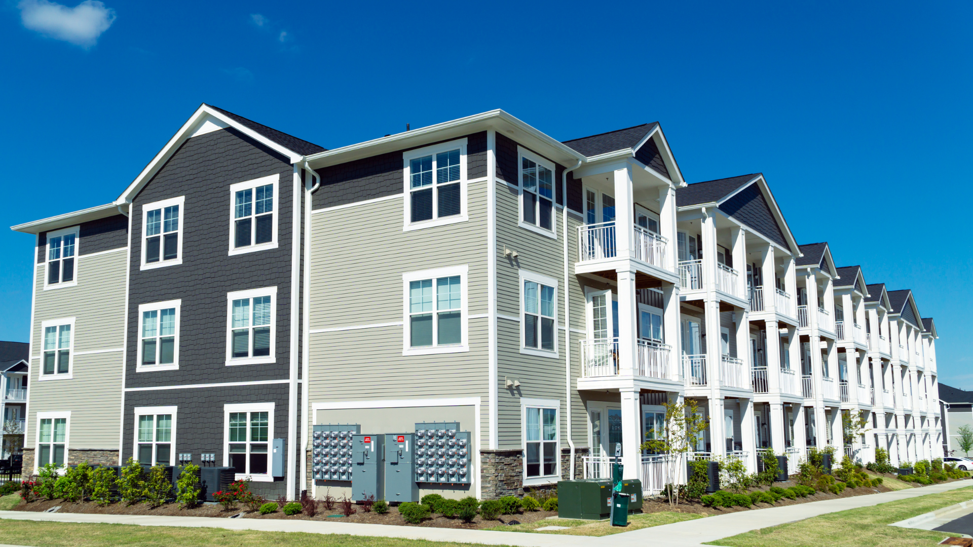 Apartment building with gray and tan siding, balconies, and bright blue sky.