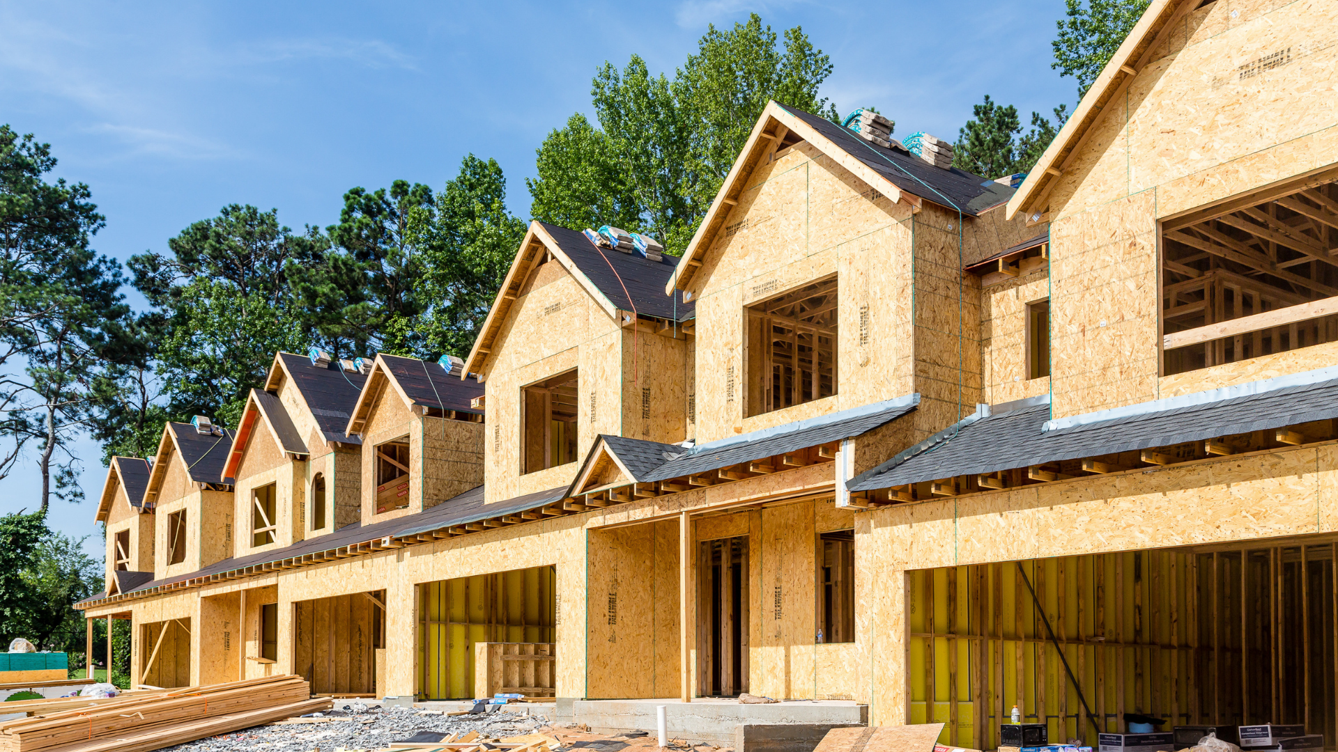 Row of townhouses under construction, framed with exposed wood, set against a backdrop of trees and a bright blue sky.