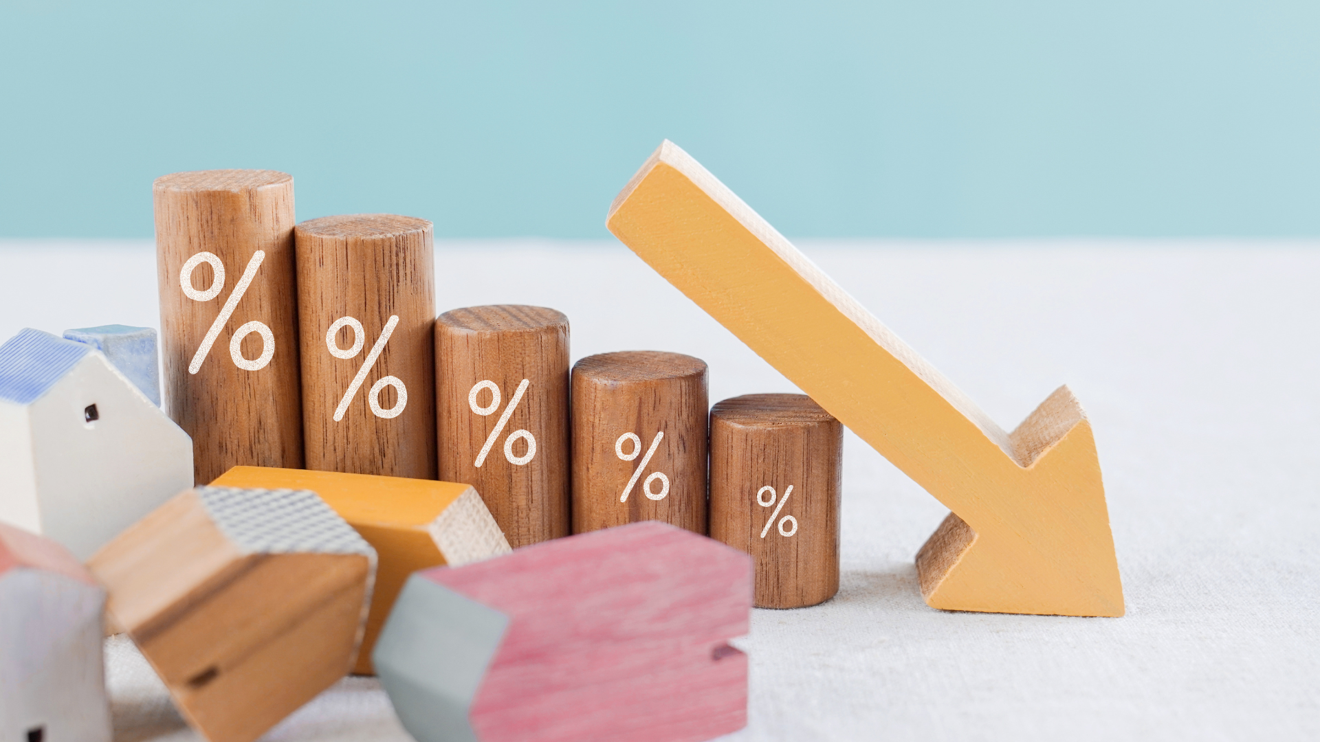 Wooden blocks with percentage signs and a downward arrow depict falling interest rates affecting small house models.