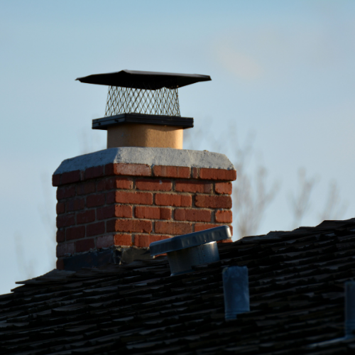 Brick chimney with a black cap on a dark-shingled roof against a blue sky.