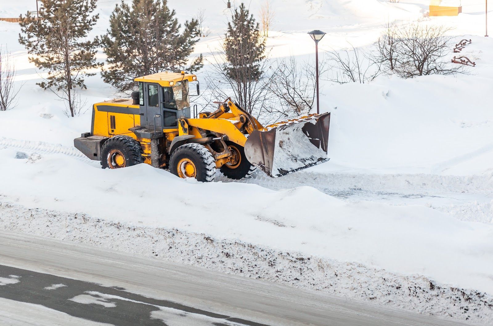 Yellow snowplow clearing snow from a road. Snowy environment with trees and a street lamp.