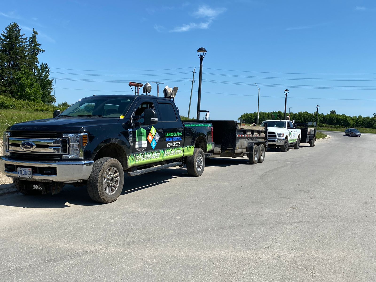 Black truck with trailer, parked on a paved road next to a white truck with a trailer, blue sky.