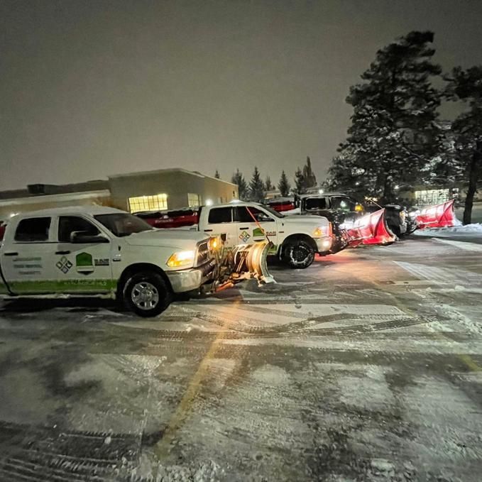 Snow plows clearing a parking lot at night. White trucks with plows; buildings and trees in background.