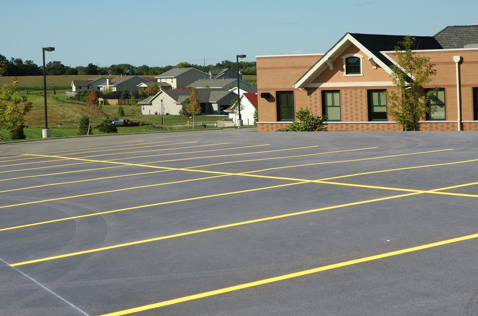 Empty parking lot with yellow lines, brick building in the background, residential area visible beyond.