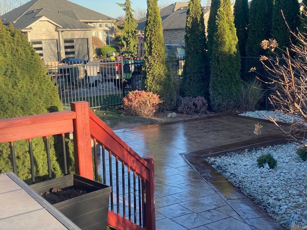 Red deck with steps leads to a wet stone patio, yard, and houses. Trees and shrubbery surround.