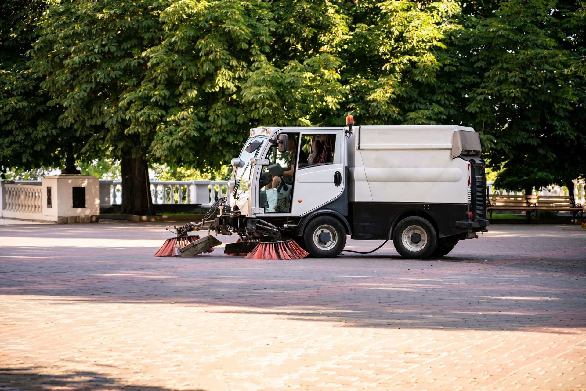 Street sweeper cleaning a paved area near a tree-lined park.