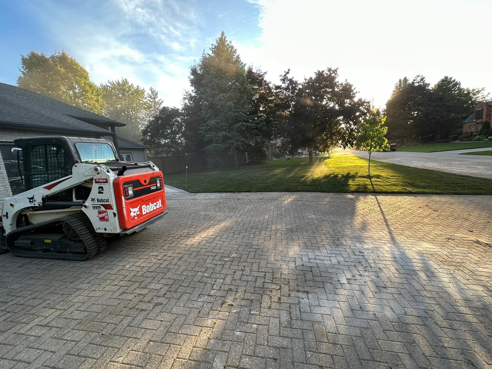 Bobcat skid-steer tractor on brick driveway; setting sun, trees, and house in background.