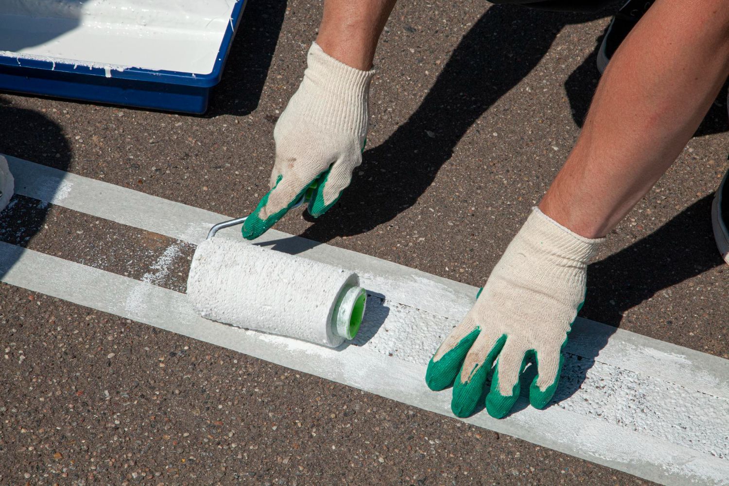 Person painting white lines on asphalt with a paint roller, wearing gloves; blue paint tray in the background.
